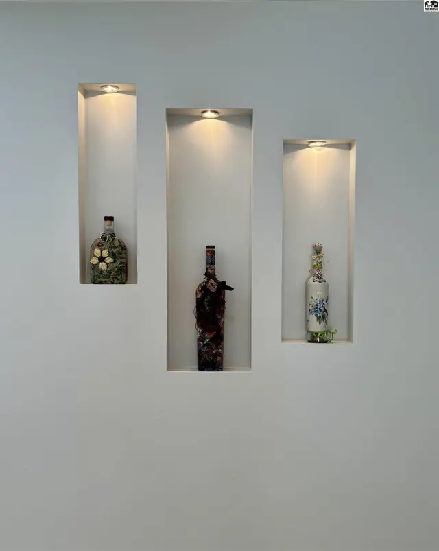 Three recessed wall niches with integrated lighting display decorative bottles and floral arrangements.