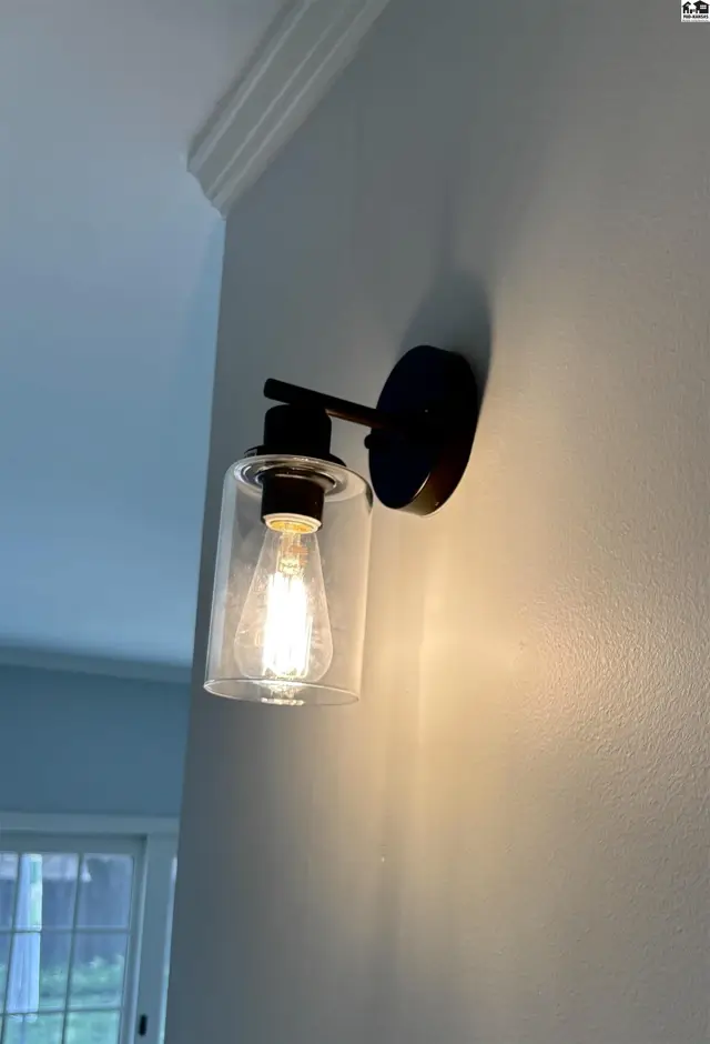 Modern black wall sconce with clear glass shade illuminates a textured white wall.