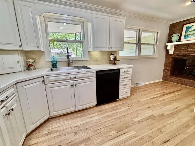 Bright kitchen with white cabinets, quartz countertops, stainless steel sink, and hardwood floors, adjacent to a brick fireplace.