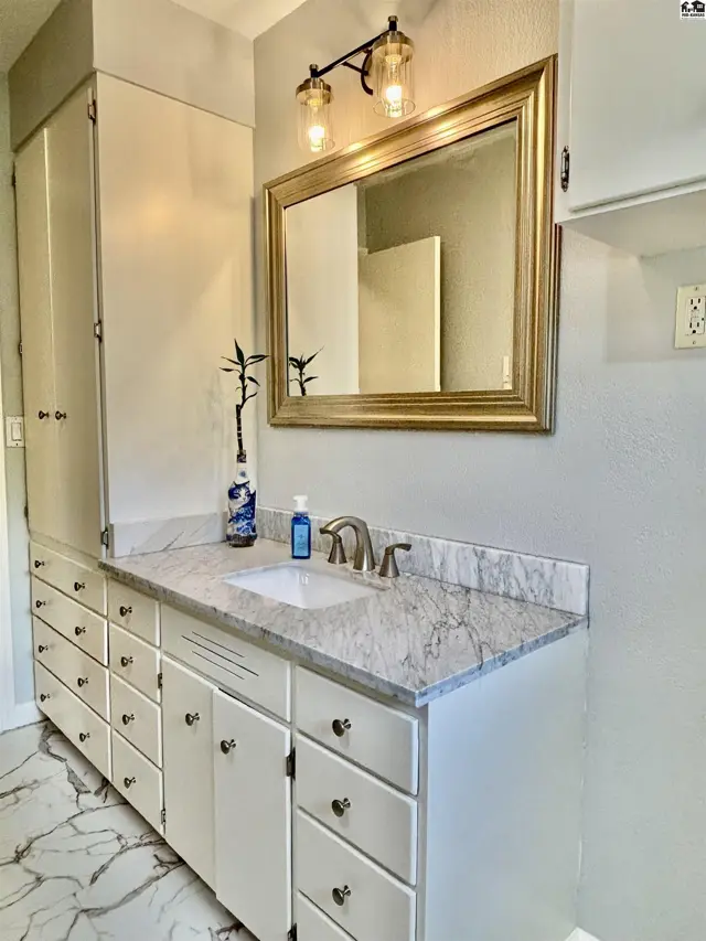 Bathroom vanity with white cabinets, marble countertop, and gold-framed mirror above a sink.