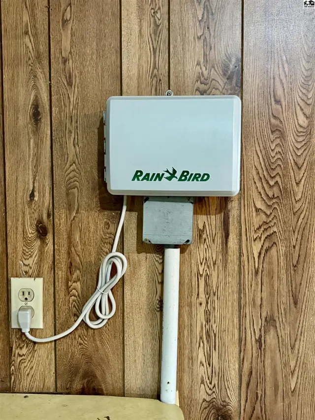 Rain Bird irrigation controller mounted on wood-paneled wall with electrical outlet and conduit.