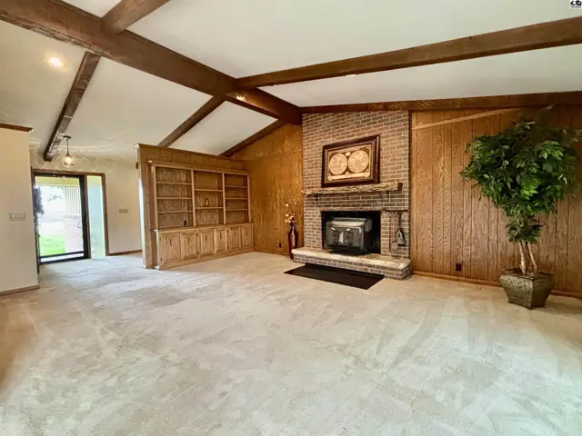 Spacious living room with vaulted beamed ceiling, brick fireplace, built-in bookshelves, and wood-paneled walls.
