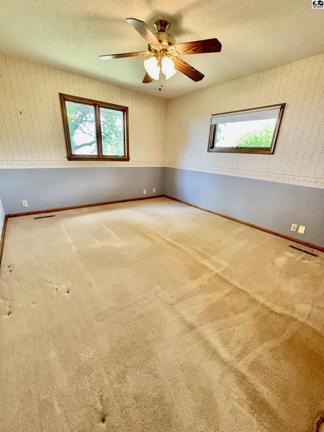 Spacious bedroom with textured wallpaper, dual-tone walls, carpet flooring, and a ceiling fan.