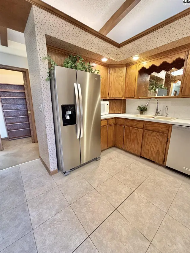 Stainless steel refrigerator in a kitchen with oak cabinets, tiled floor, and patterned wallpaper.