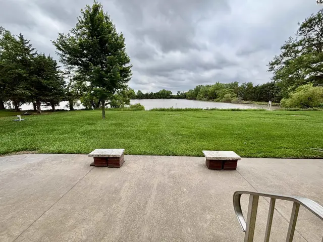Concrete patio with two stone-topped benches overlooks a serene lake surrounded by lush green trees.