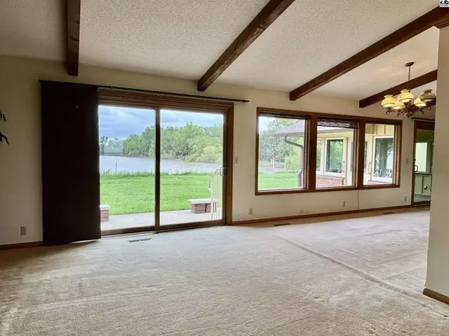 Spacious living room with vaulted beamed ceiling, large windows, and carpeted floors overlooking a serene waterfront.