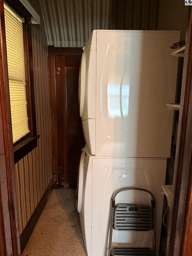 Stacked white washer and dryer in a narrow laundry closet with striped wallpaper and wood trim.
