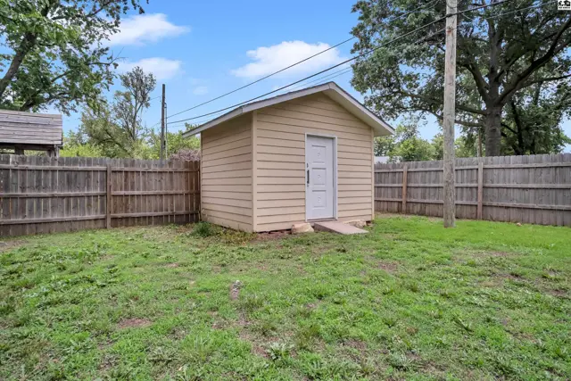 Tan shed with white door sits in a fenced backyard with green grass and mature trees.