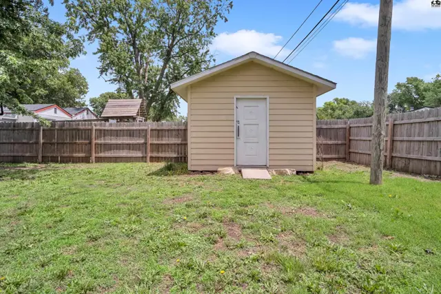 Small tan shed with white door sits in a fenced backyard with green grass.