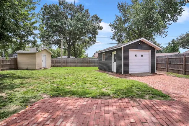 Backyard with a detached dark gray garage, tan shed, and red brick patio.