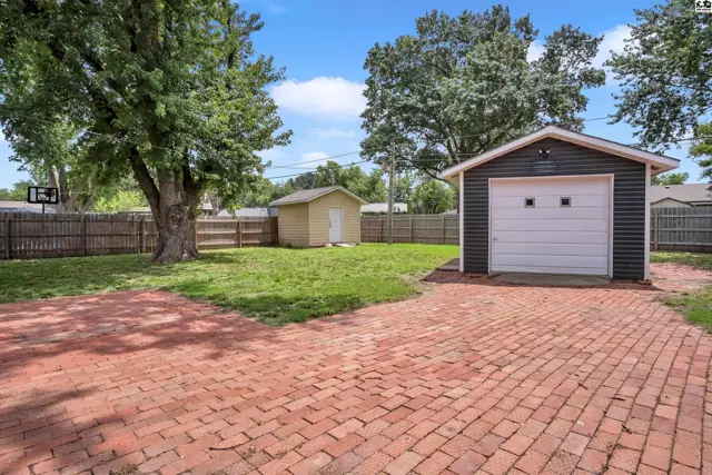 Brick driveway leads to a dark-sided garage with a white door, next to a grassy yard and shed.