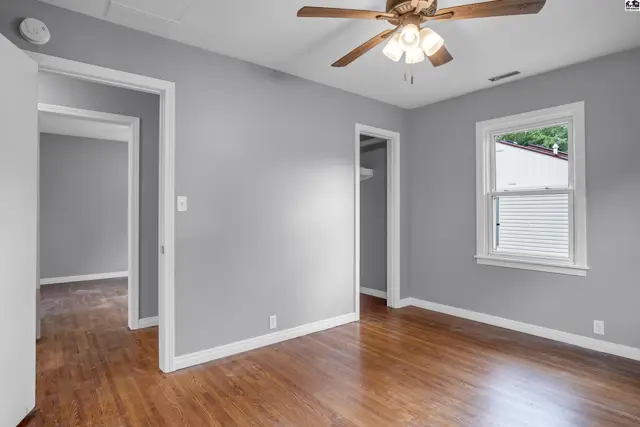 Spacious room with gray walls, hardwood floors, and a ceiling fan, opening to other rooms.