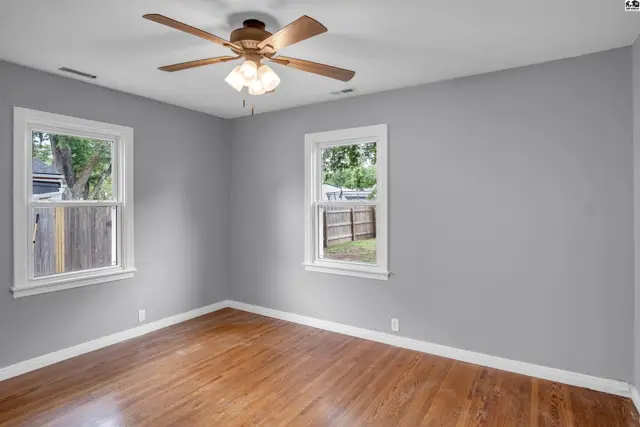 Bright bedroom with neutral gray walls, hardwood floors, and two windows overlooking a fenced yard.