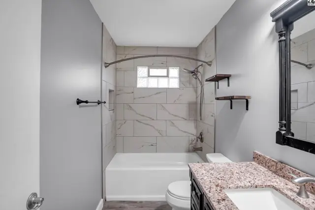 Modern bathroom with white tub, marble tile shower, granite vanity, and dark wood cabinets.