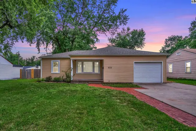 Tan siding ranch home with a brick walkway, garage, and mature trees under a colorful sunset sky.