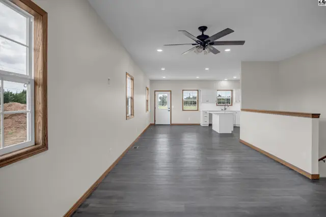 Spacious living area with dark wood-look flooring, white walls, and a ceiling fan, opening to a kitchen.