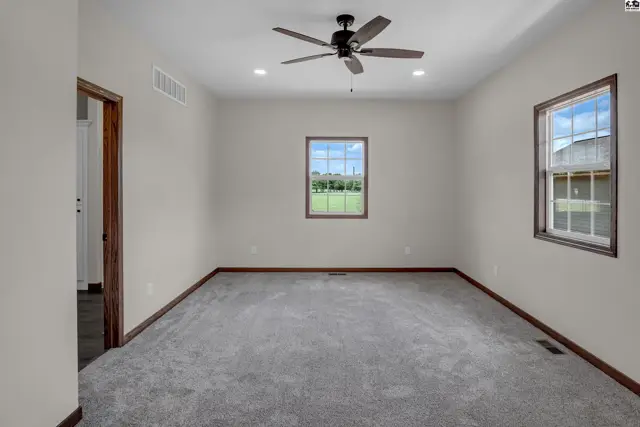Spacious bedroom with plush gray carpet, neutral walls, ceiling fan, and large windows overlooking a green field.