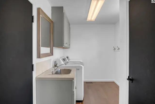 Bright laundry room with white washer/dryer, stainless sink, and gray cabinetry.