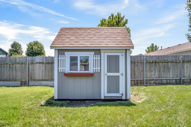 A charming gray shed with white trim, shutters, and a brown shingled roof sits in a grassy yard.