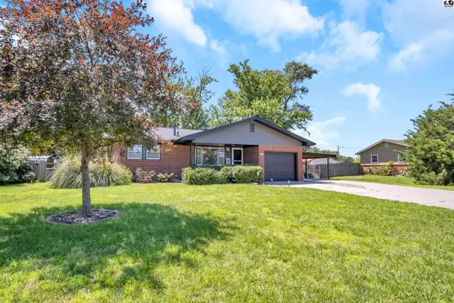 Brick ranch home with a covered carport, mature trees, and a well-maintained lawn under a blue sky.