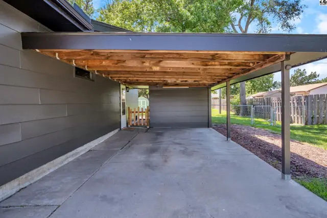 Carport with exposed wooden beams, gray siding, and concrete driveway.