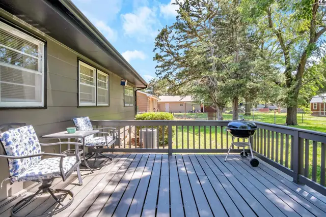 Spacious wooden deck with seating and grill overlooks a sunny backyard with mature trees.
