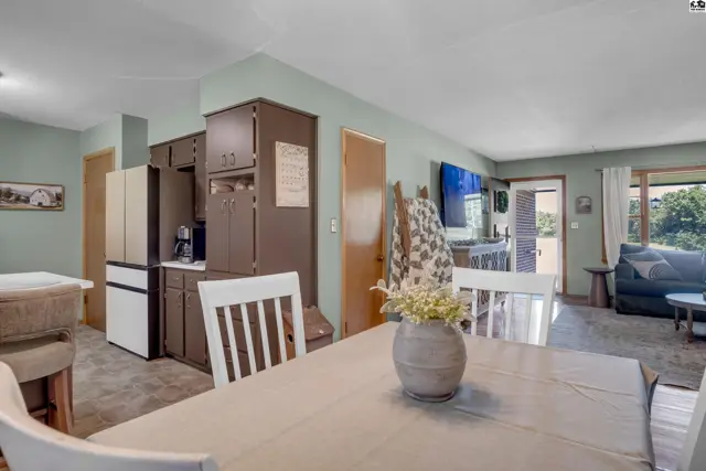 Dining area and kitchen with white chairs, brown cabinets, and a large window overlooking a living room.