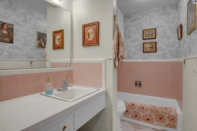 A bathroom with pink tile wainscoting, floral wallpaper, and a white vanity with a sink.