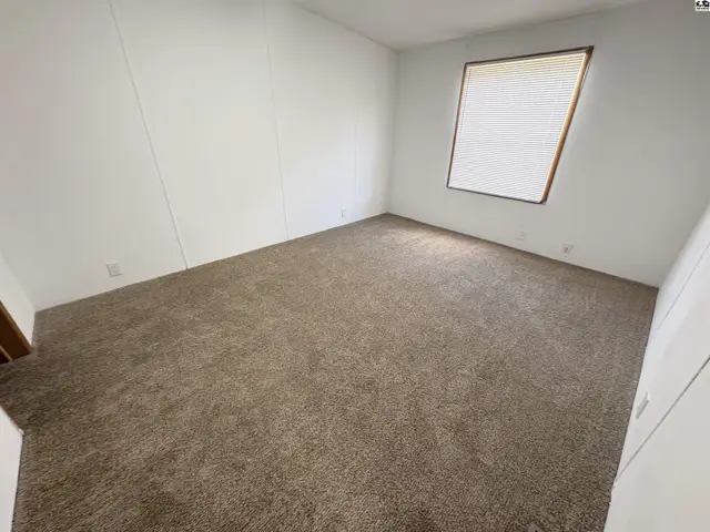 Empty bedroom with neutral carpet, white paneled walls, and a large window with blinds.