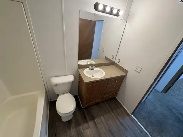 Bright bathroom with white tub, toilet, and vanity featuring wood cabinets and laminate flooring.