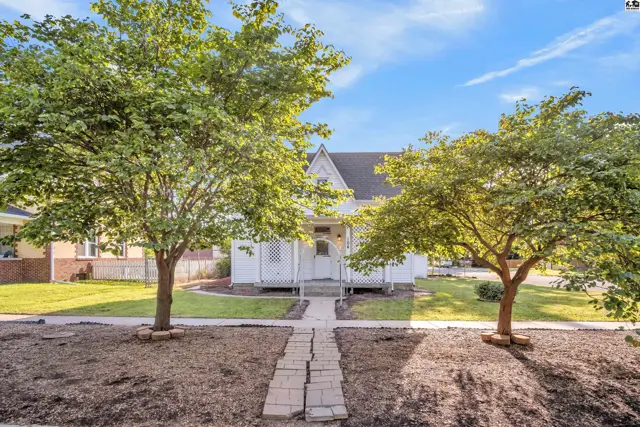 Charming white clapboard house with a covered porch, flanked by mature trees and a brick walkway.