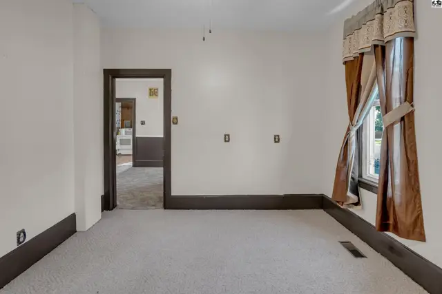 Spacious room with neutral carpet, dark trim, and brown satin curtains framing a window.