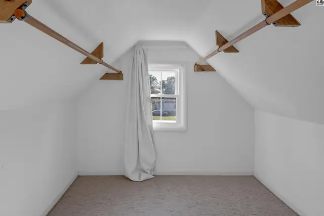 Attic room with sloped ceilings, a single window with white curtains, and carpeted floor.