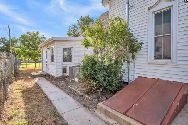 White siding exterior with a concrete walkway, metal cellar doors, and mature landscaping.