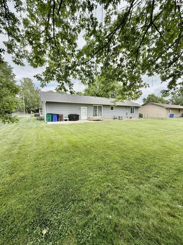 Backyard with a large green lawn, patio, and gray siding house under leafy trees.