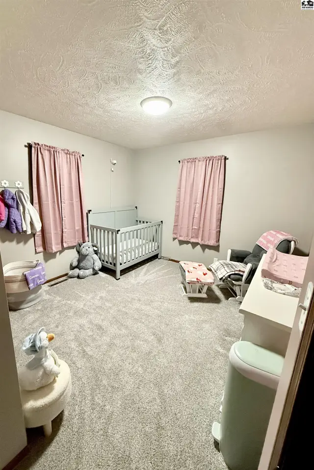 Nursery with gray crib, plush toys, pink curtains, and a dresser on plush gray carpet.