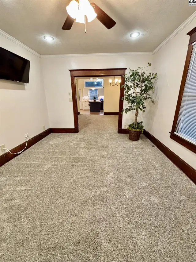 Living room with plush gray carpet, dark wood trim, and a view into the kitchen.