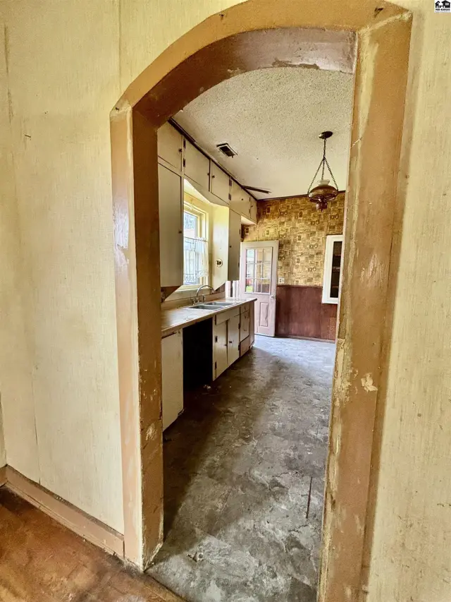 Arched doorway reveals a vintage kitchen with worn cabinets, textured wallpaper, and concrete floors.