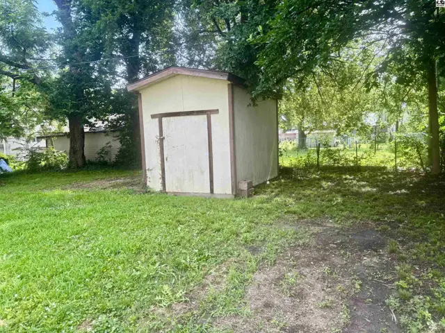 A small, light-colored shed with a wooden door sits in a grassy yard surrounded by trees.
