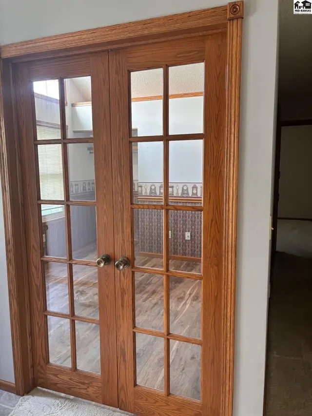 Oak double doors with glass panes open to a room with patterned wallpaper and wood-look flooring.