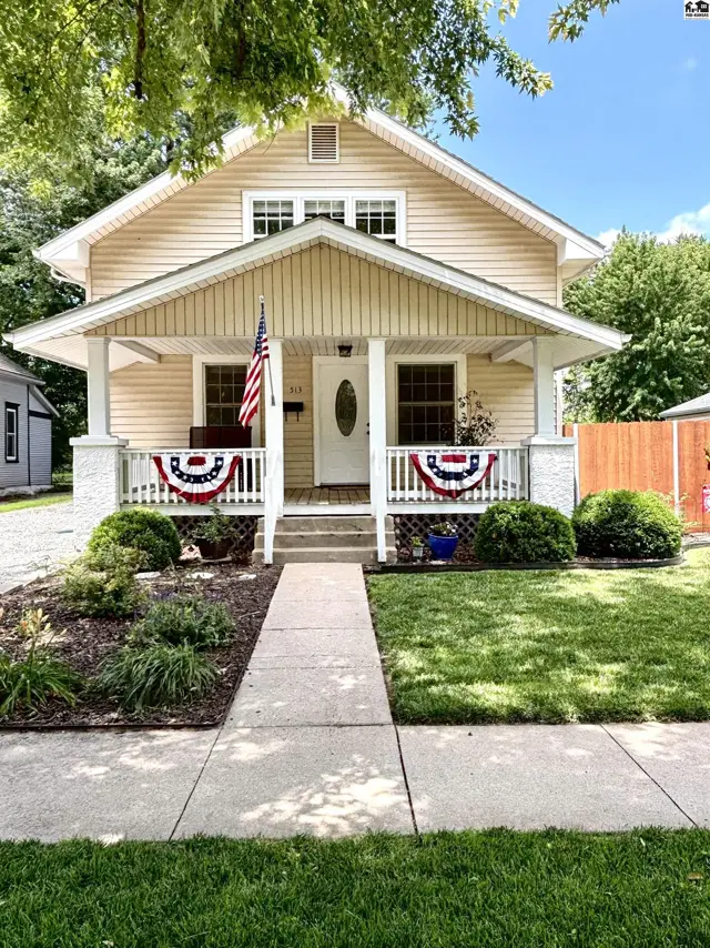 Charming tan bungalow with a welcoming covered porch, white railings, and lush landscaping.