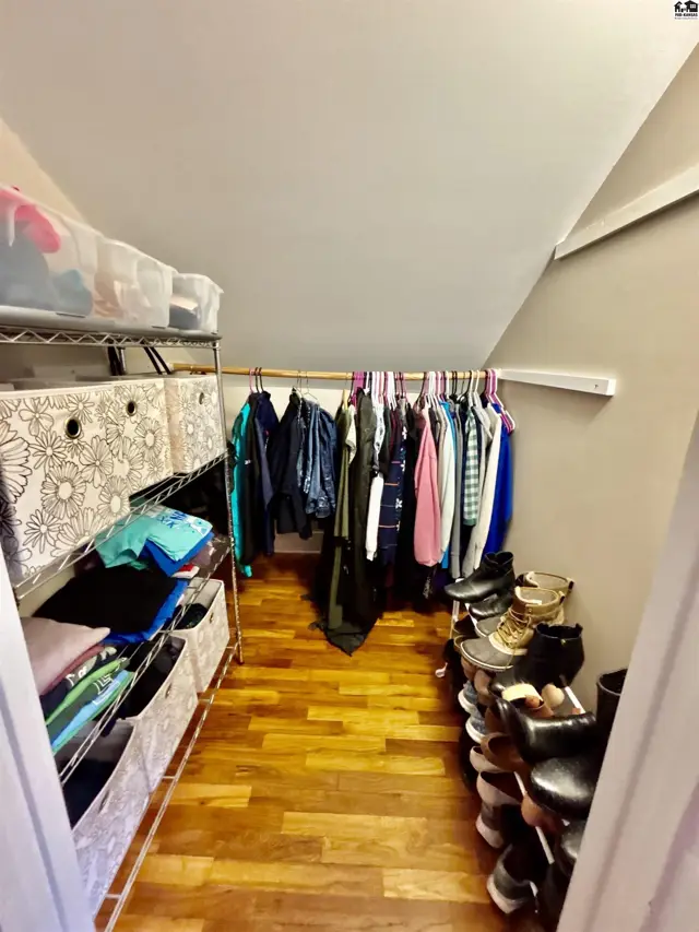 Sloped-ceiling walk-in closet with wood floors, open shelving, and a rod displaying colorful clothing.