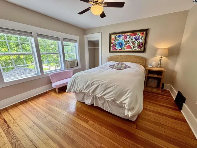 Cozy bedroom with hardwood floors, large windows, a ceiling fan, and a colorful floral art piece.