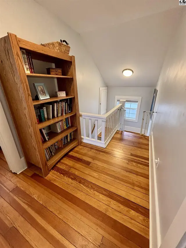 Warm wood floors and a tall bookshelf line this bright, attic-level landing with a window and railing.