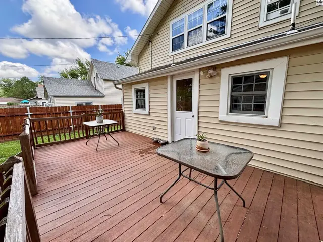 Spacious wooden deck with glass-top tables and a fenced yard, adjacent to a light-colored siding house.