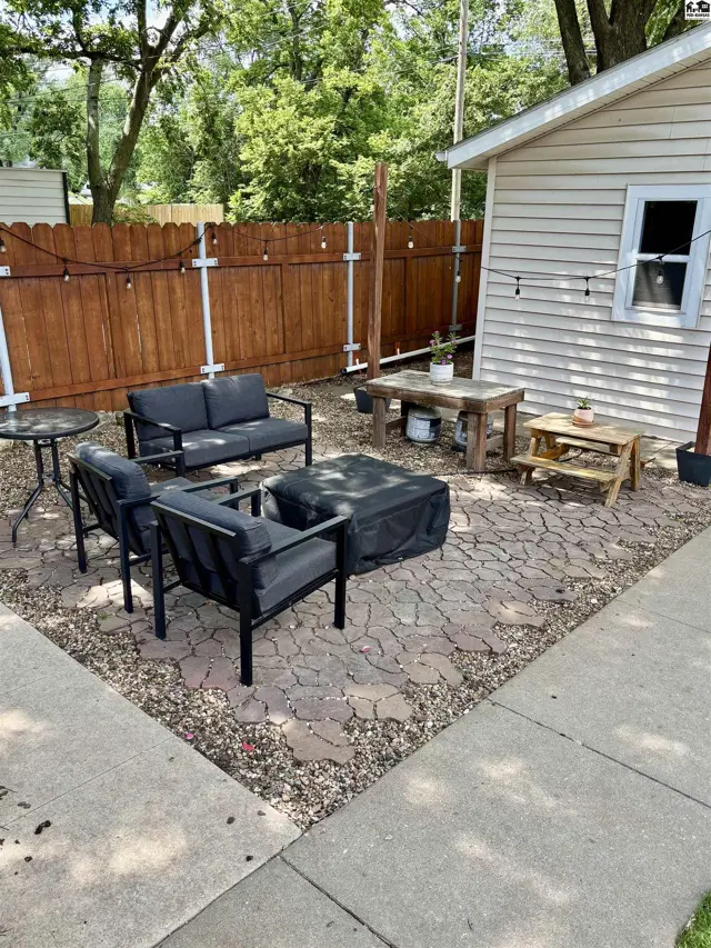 Outdoor patio with stone pavers, gravel, seating, and wooden tables next to a light-colored shed.