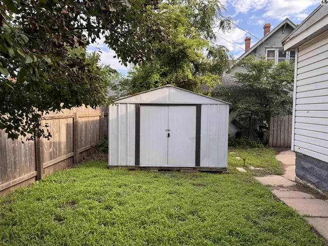 White metal shed with double doors sits in a grassy backyard with a wooden fence.