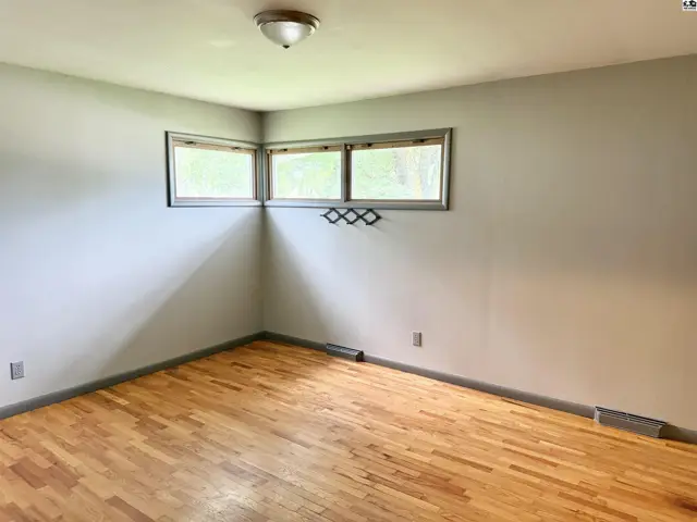 Corner of a room with light gray walls, three windows, and polished hardwood floors.