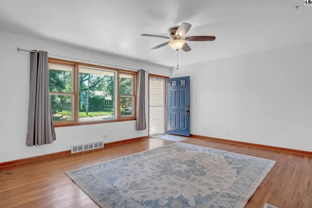 Bright living room with hardwood floors, large windows, a ceiling fan, and a blue front door.