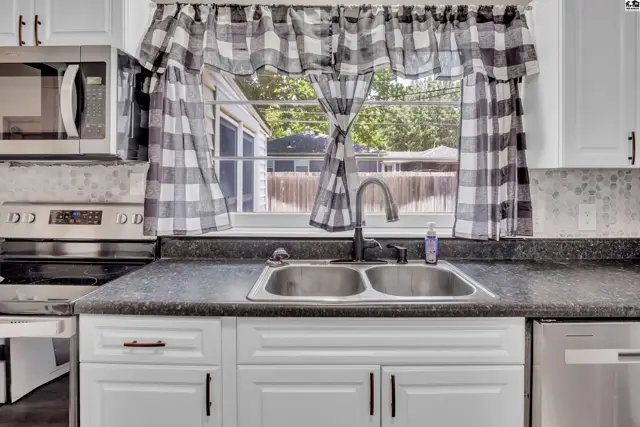 Kitchen with white cabinets, granite countertops, double sink, and checkered curtains framing a backyard view.